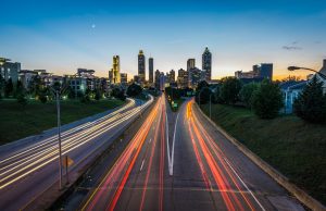 A long-exposure photo of a city highway at dusk shows light trails from cars heading toward the downtown skyline near PI Agency Atlanta, with greenery on both sides and a small crescent moon illuminating the tall buildings.