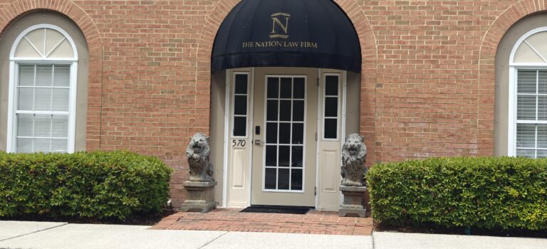 The entrance to The Nation Law Firm features a black awning, double glass doors, two stone lion statues on either side, symmetrically trimmed bushes, and arched windows on a red brick building.