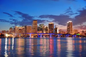A vibrant FL city skyline at dusk with tall buildings lit up, a brightly illuminated bridge, and colorful reflections shimmering on the water in the foreground—capturing the energetic spirit of PI Agency Orlando.
