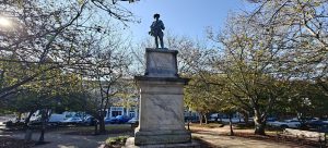 A stone statue of a soldier stands atop a large pedestal in a park surrounded by trees with autumn leaves. Cars are parked on the street in the background under a clear blue sky.
